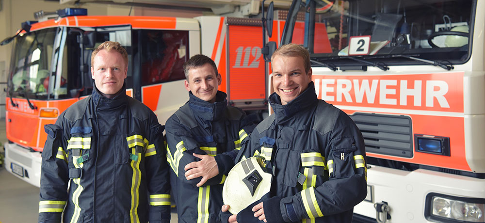Drei Feuerwehrmänner stehen vor den Feuerwerkautos und lächeln in die Kamera.