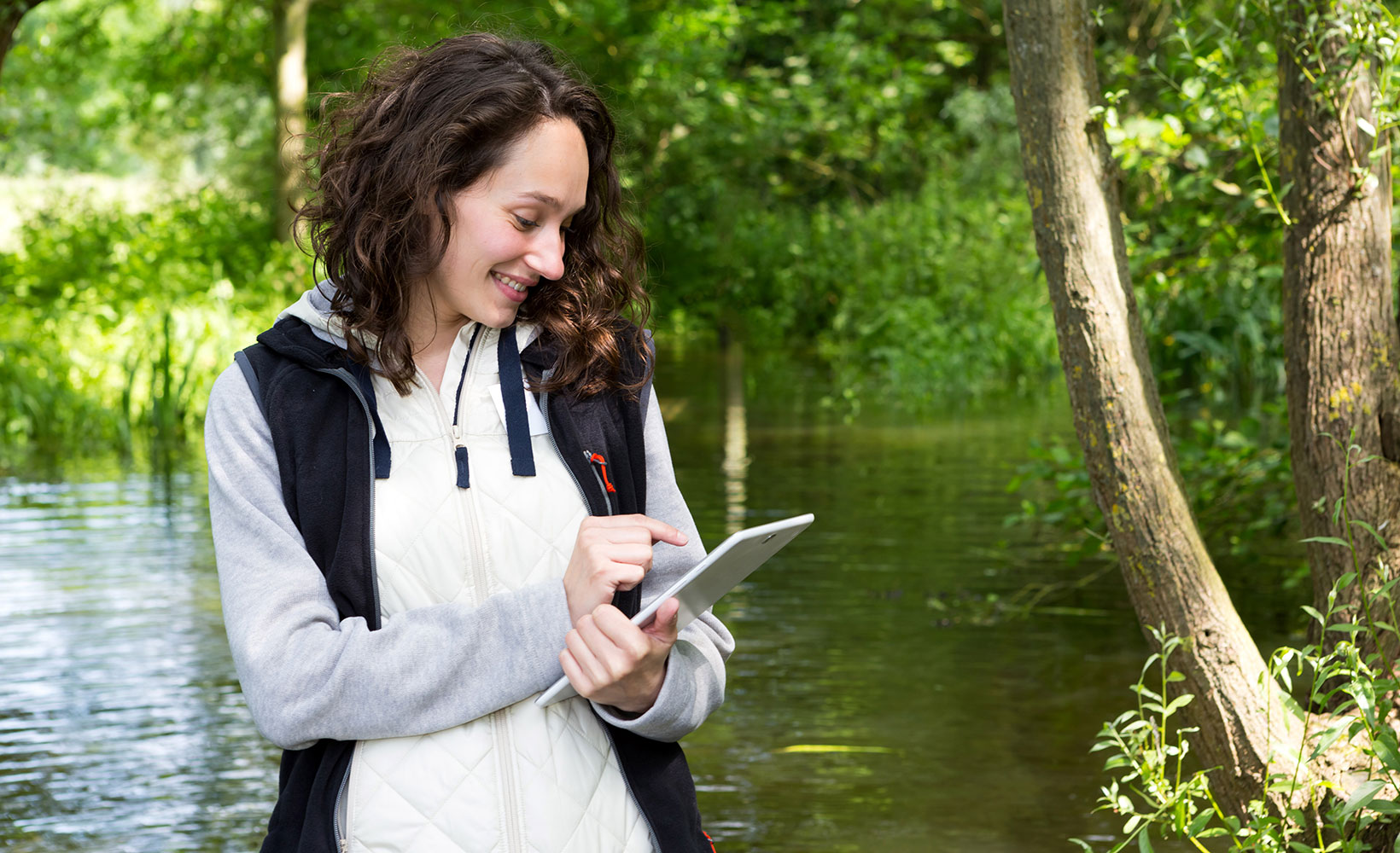 Eine Wasserbäuerin tippt lächelnd etwas auf ihrem Tablet. Sie steht vor einem Fluss mitten in der Natur.