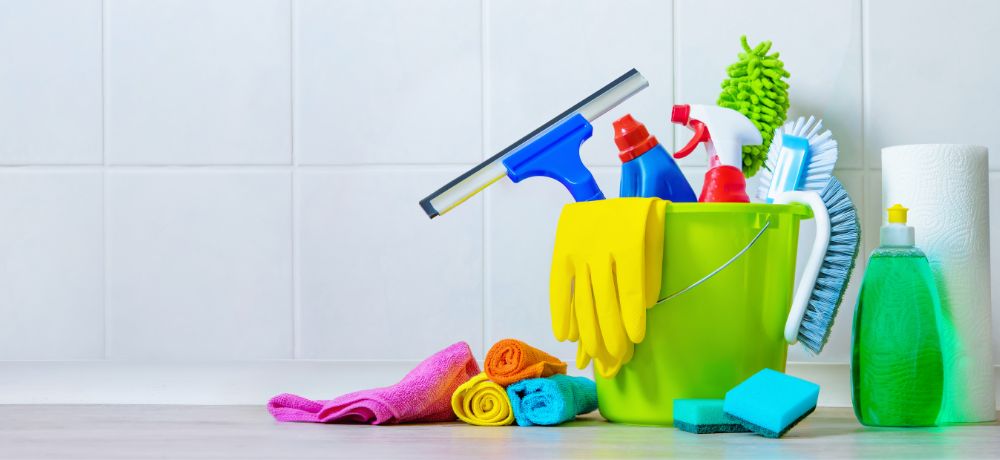 Vor einer weiß, gefliesten Wand steht rechts ein grüner Eimer mit verschiedensten Putzwerkzeugen und Putzmitteln.