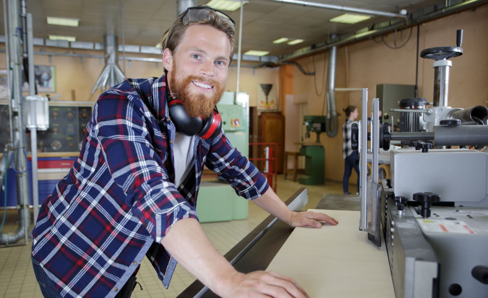 Holzmechaniker trägt seinen Ohrenschutz um den Hals, steht vor einer Maschine und lächelt in die Kamera. Er steht auf der linken Seite des Bildes und lehnt sich auf der rechten an einen Tisch. Auf diesem befindet sich eine Maschine.