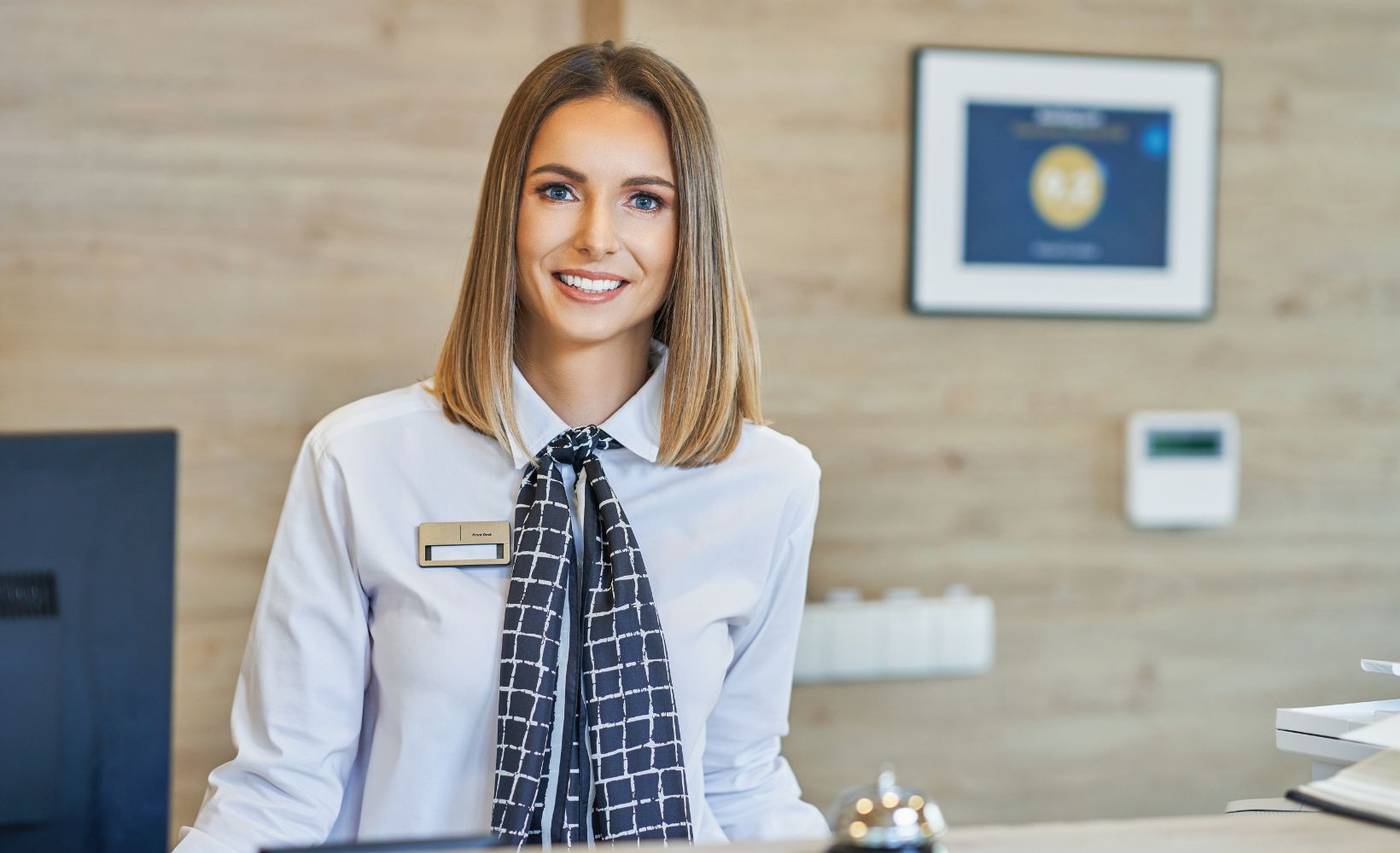 Eine ausgelernte Hotelfachfrau in ihrer Uniform lächelt über die Rezeption in die Kamera.