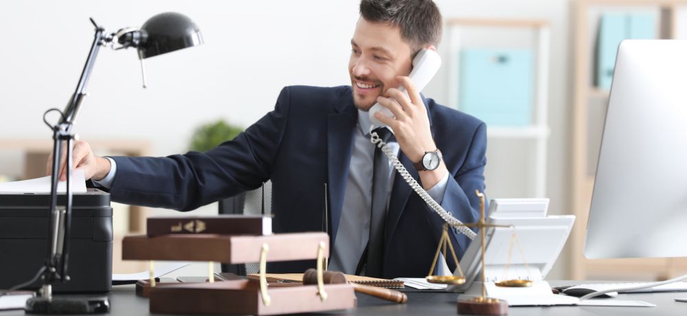 Ein Rechtsanwaltsfachangestellter sitzt an seinem Schreibtisch in seinen Anzug, während er ein Klienten am Telefon hat und Unterlagen aus dem Drucker nimmt.