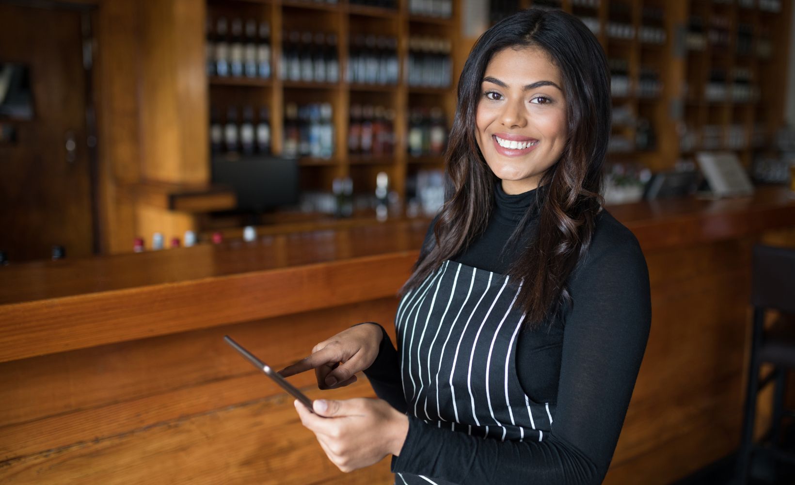Eine Restaurantfachangestellte in einer schwarz und weiß gestreiften Schürze hält ein Tablet in ihrer Hand während sie was antippt und in die Kamera lächelnd.