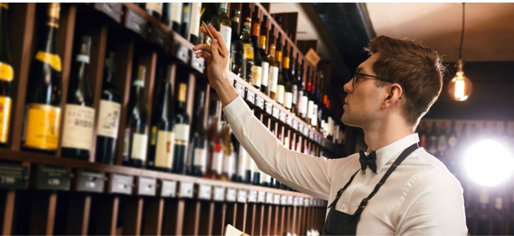 Ein Restaurantfachangestellter steht vor einem riesen Wandregal voller verschiedener Weinflaschen, gekleidet in einem weißen Hemd mit schwarzer Schleife und schwarzer Schürze. Er streckt eine Hand zu einer Weinflasche aus und in der anderen hält er ein Notizheftchen.