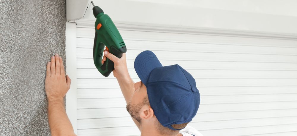 Ein Rollladen und Sonnenschutzmechatroniker schraubt eine Befestigung für das Fenster mit einer Bohrmaschine an.