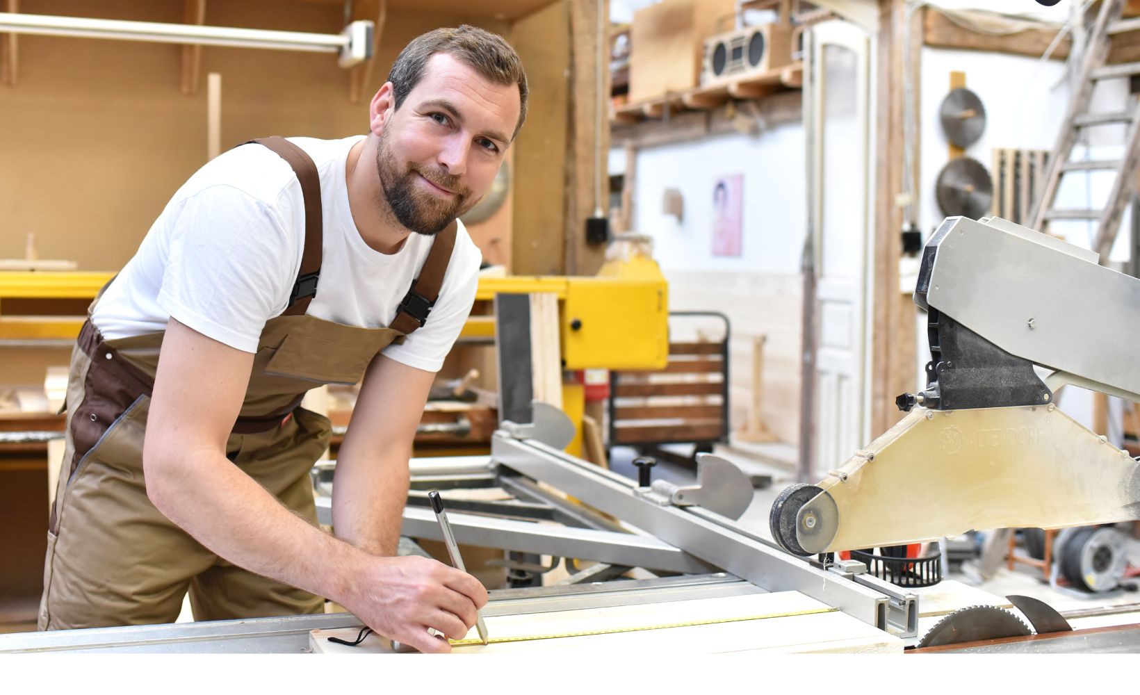 Ein Mann schaut lächelnd in die Kamera währenddessen er Holz an der Kreissäge ausmisst.