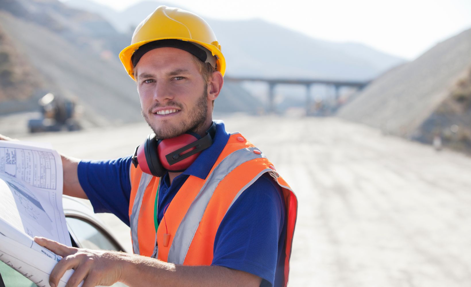 Ein Straßenbauer steht mit Bauplan und Arbeitskleidung auf einer Baustelle.