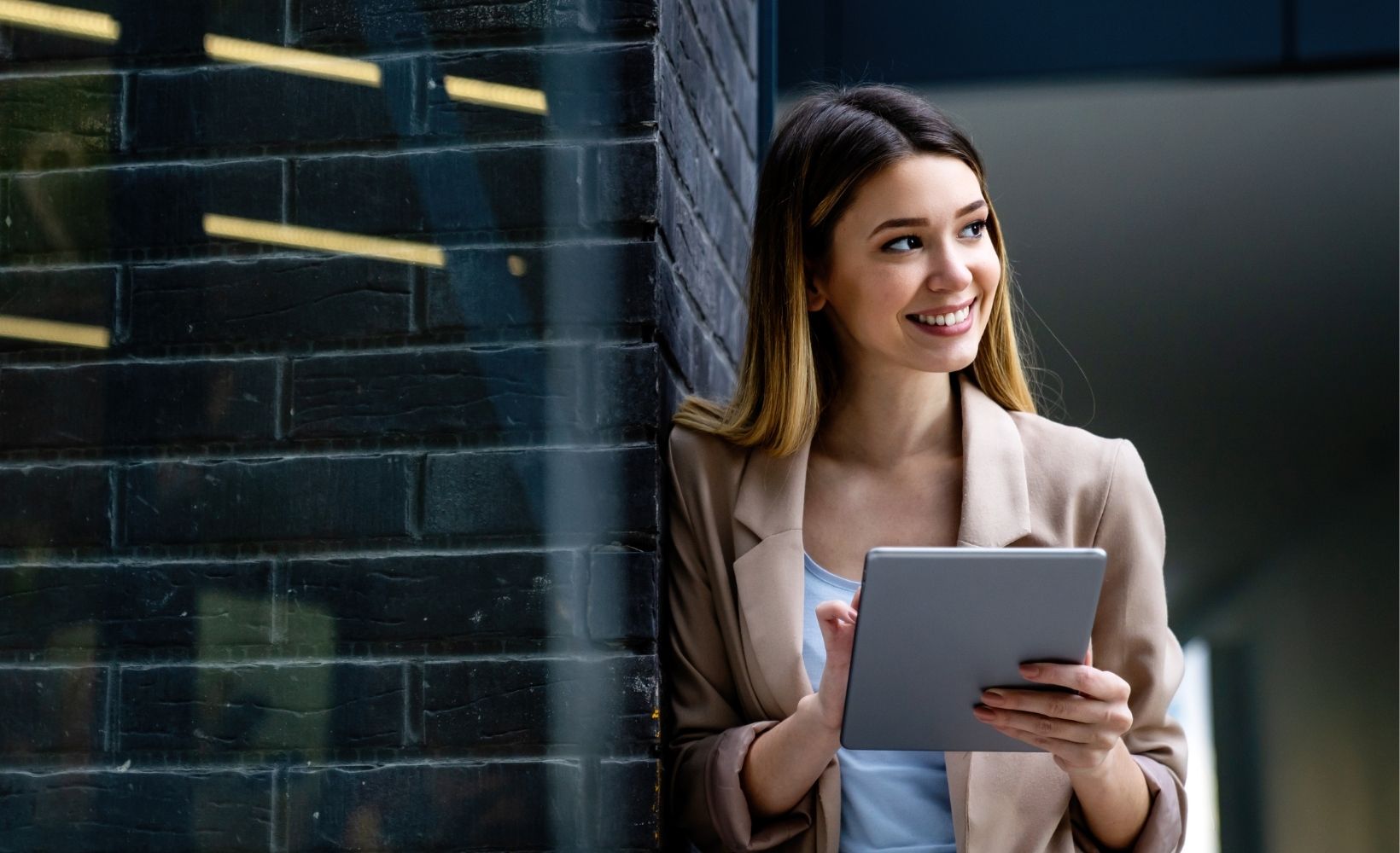 Lächelnde Frau an einer Mauer angelegt. In der Hand ein Tablet, welches Sie bedient.