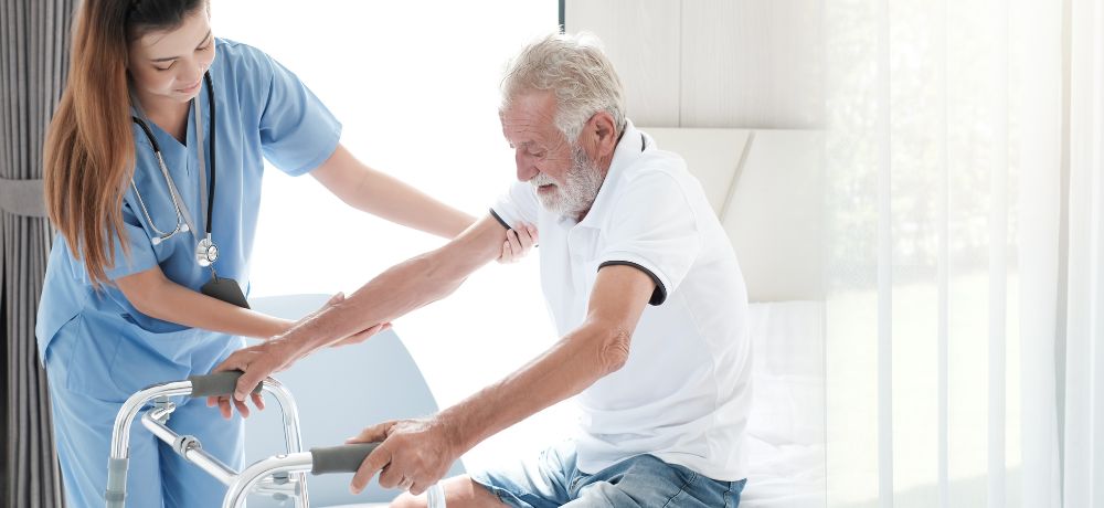 Altenpflegerin hilft einem älteren Mann mit Rollator aufzustehen.