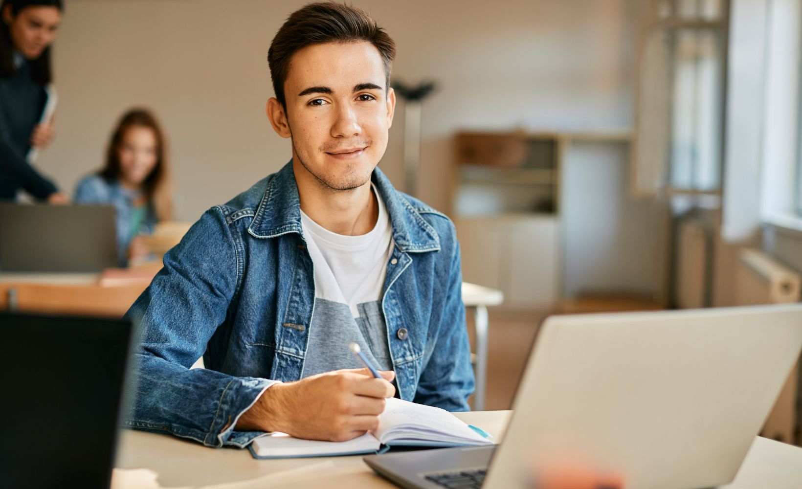 Dualer Student sitzt im Klassenzimmer am Laptop und lächelt in die Kamera.