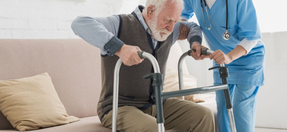 Ein Pflegeassistent hilft einem älteren Mann mit Gehhilfe aufzustehen.