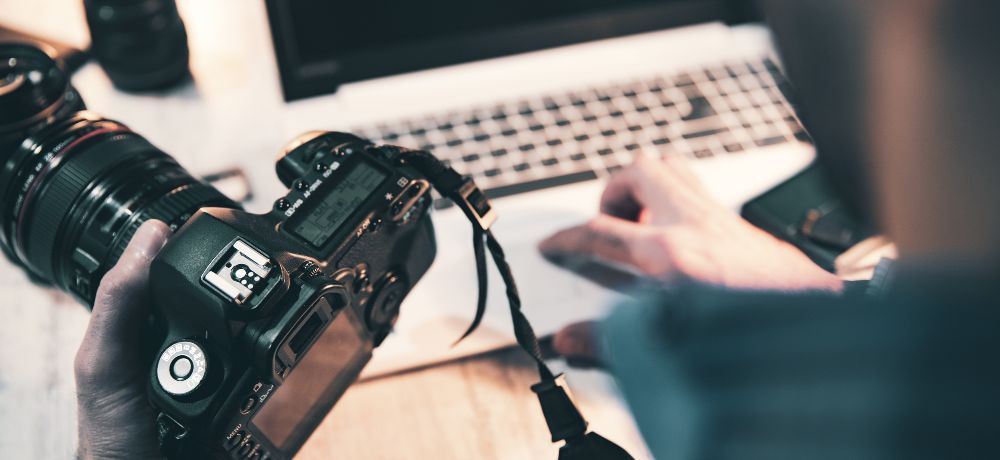 Foto-und medientechnische Assistentin hält eine Kamera in der Hand und arbeitet am Laptop.