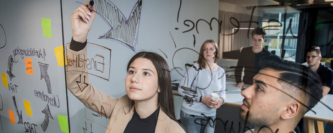 Mehrere junge Menschen arbeiten innerhalb eines Workshops zusammen und malen ein Schaubild auf eine Glaswand.