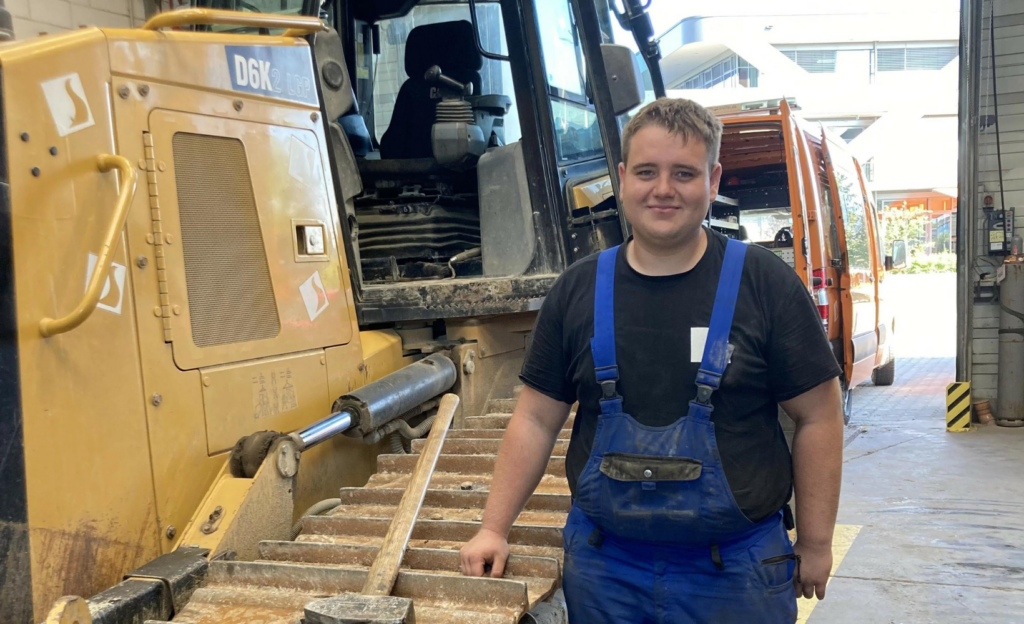 Baumaschinenmechatroniker-Azubi Justin Bareiß neben einem Baustellenfahrzeug in einer Halle.