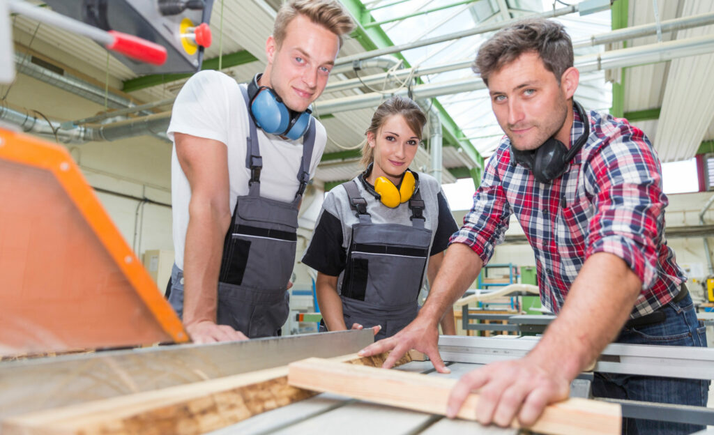 Ein Ausbildungsleiter erklärt zwei Auszubildenden in einem Schreinerbetrieb die Arbeit mit Holz. Zum Jahresstart 2024 steigt der Mindestlohn für Azubis.