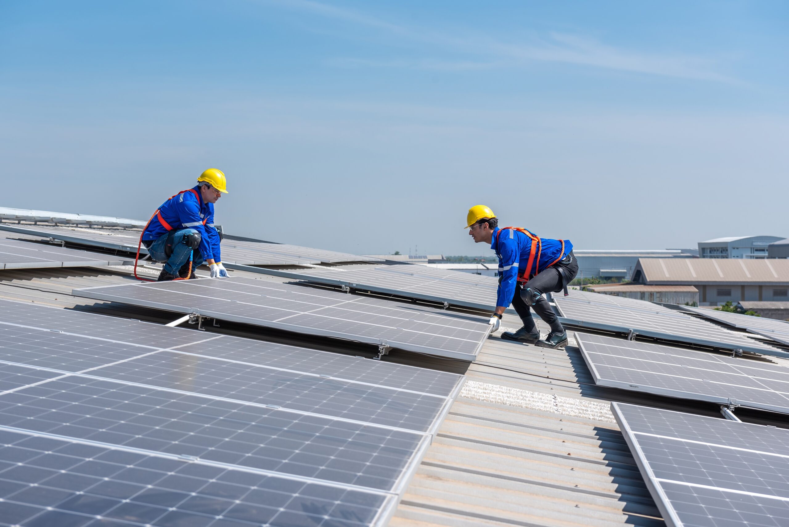 Two solar panel technicians in hard hats and safety gear install solar panels on a commercial building