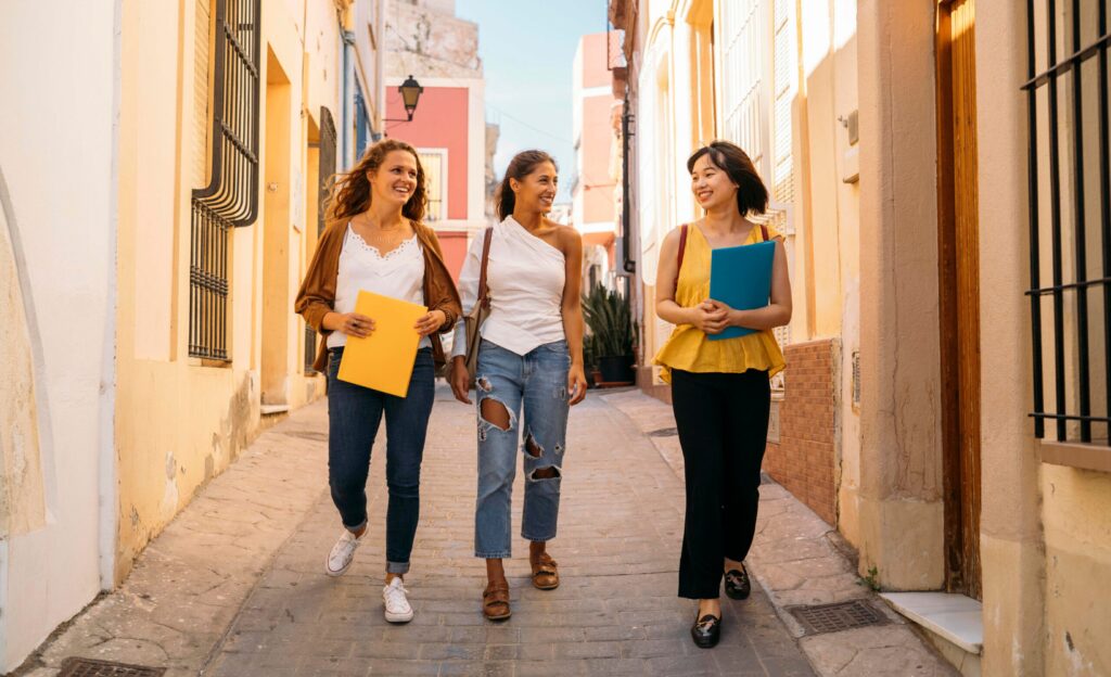 Drei Frauen laufen während ihres Gap Years nebeneinander im Sonnenschein durch eine schattige Gasse.