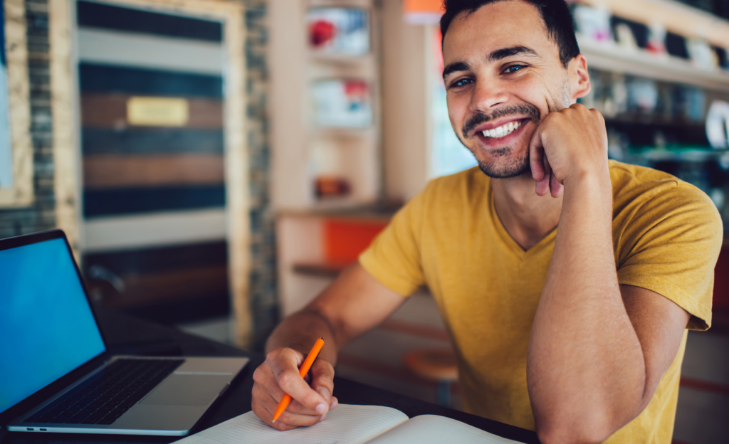 Lachender Student sitzend an einem Schreibtisch mit Heft und Laptop und einem Stift in der Hand.