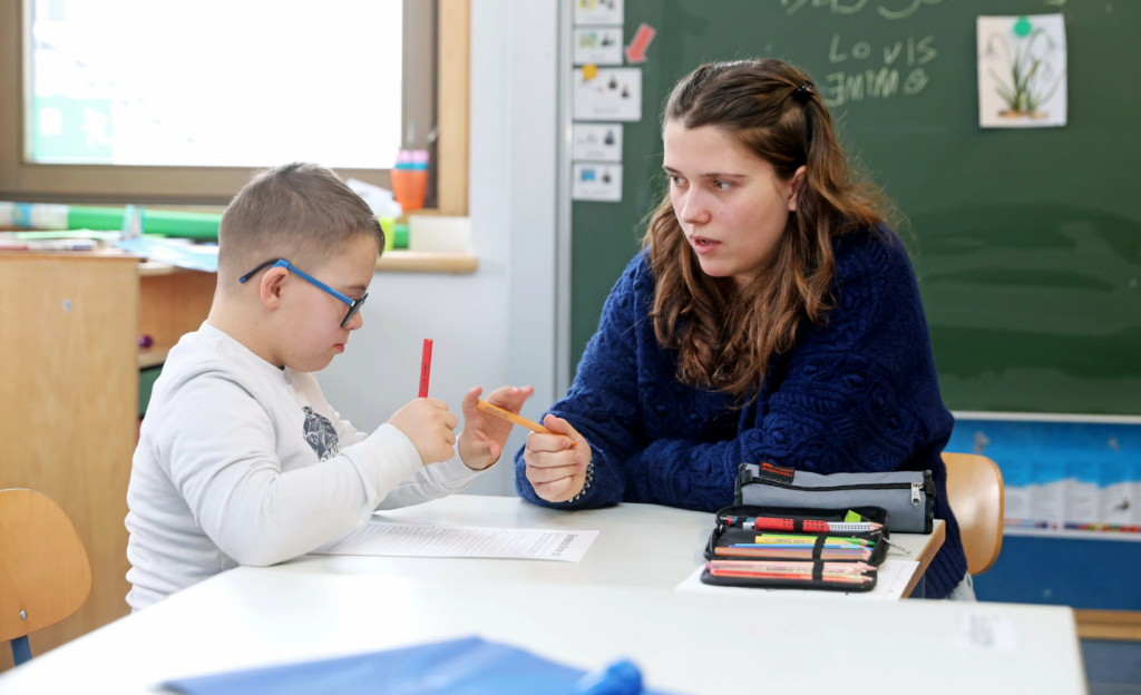 Lena nun, die ein FSJ an einer Sonderschule absolviert, unterhält sich mit einem Kind, das gerade einen Stift von ihr entgegennimmt.