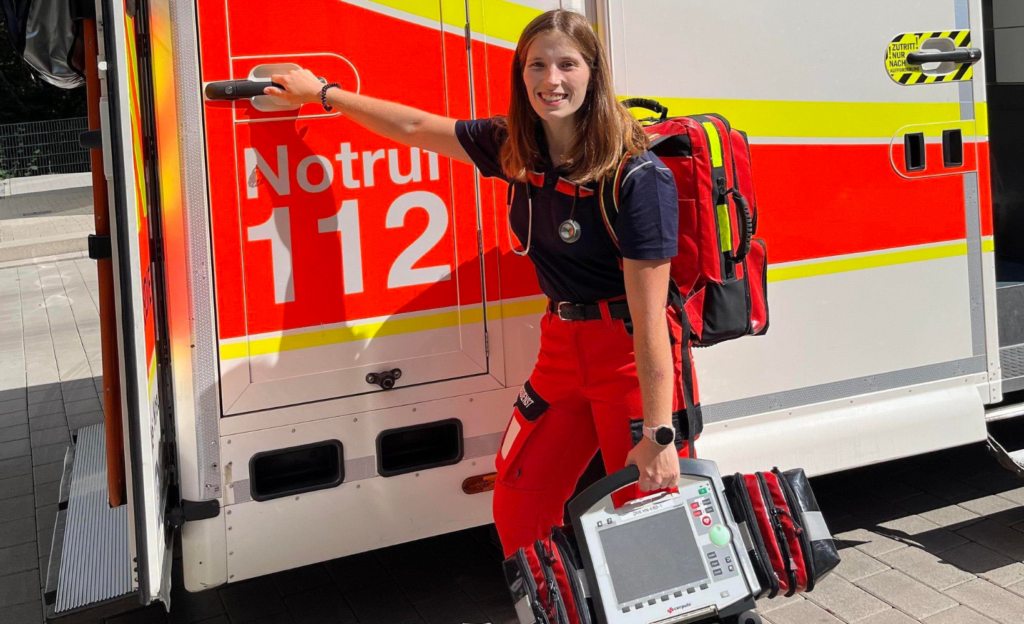 Sophia Weißschuh, die eine Ausbildung zur Notfallsanitäterin macht, steht im Sonnenschein an der Seite eines Rettungswagens und hält eine Türklinke in der Hand.