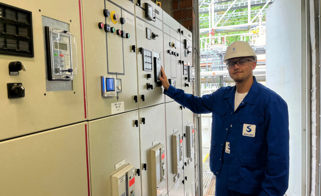 Ein Mann in blauer Arbeitskleidung und Schutzhelm steht vor einer Schalttafel mit elektrischen Steuergeräten.