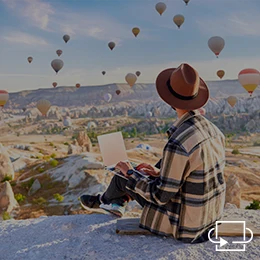 Ein Mann arbeitet im Freien am Laptop auf einer Erhöhung. Im Hintergrund fliegen viele Heißluftballons