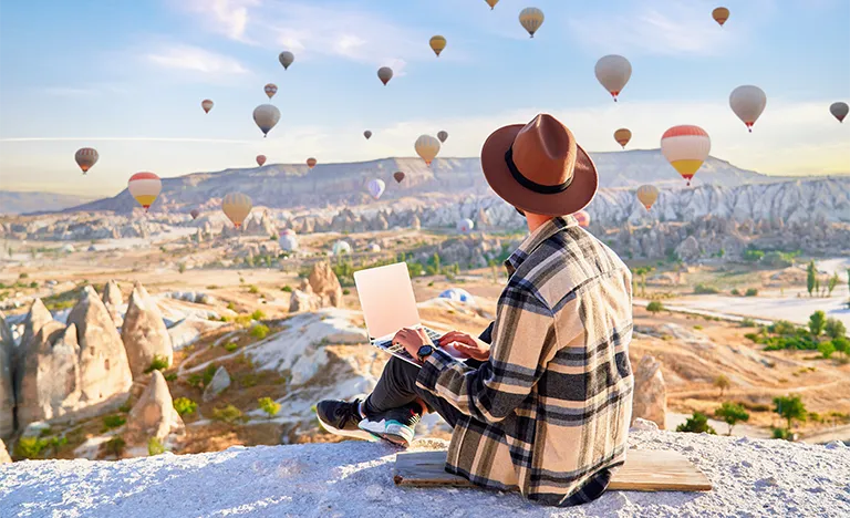Work & Travel: Ein Mann arbeitet im Freien am Laptop auf einer Erhöhung. Im Hintergrund fliegen viele Heißluftballons