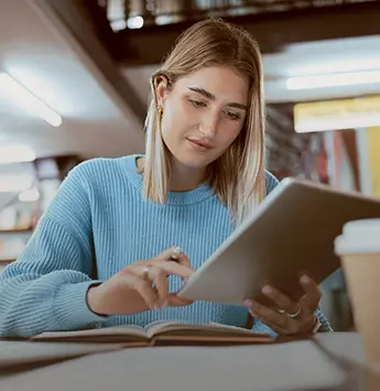 Frau in Bibliothek sitzt am Tisch und schaut auf ihr Tablet.