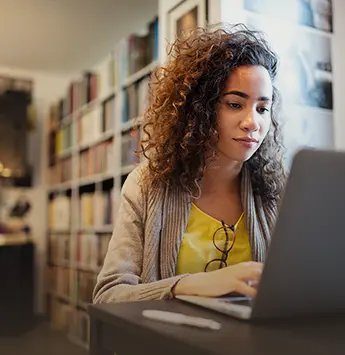 Junge Frau sitzt in der Bibliothek am Tisch und schaut dabei auf ihr Notebook.